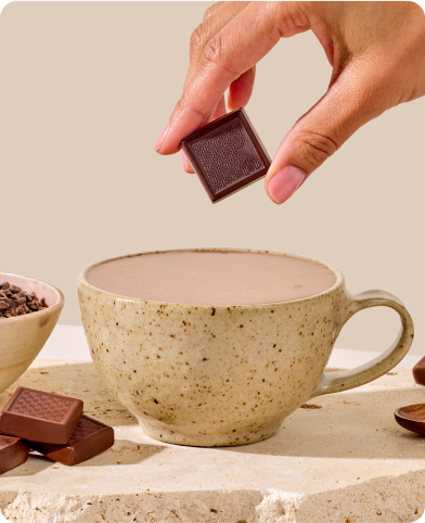 Hand holding chocolate square above a speckled cup of hot cocoa.